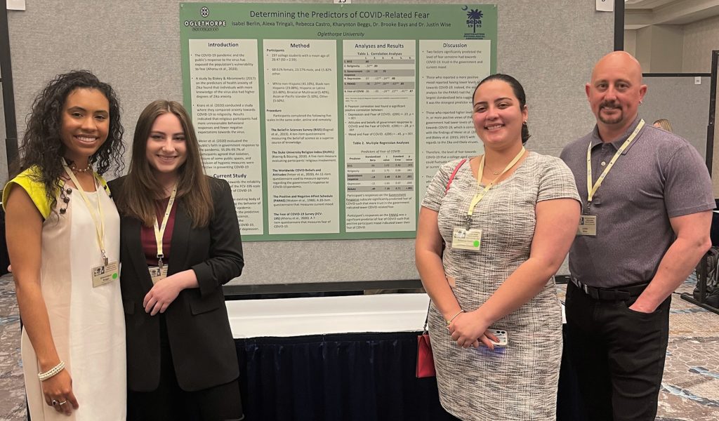 Three students and a professor stand with a presentation poster depicting their research on "Determining the Predictors of COVID-Related Fear".