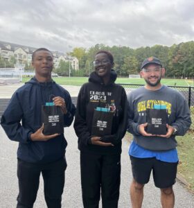 Three students standing at the turf field each holding a gift bag from The Joint Chiropractor that reads "Relief Recovery Wellness. The Joint Chiropractor."