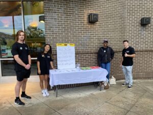 Four students standing next to a table set up in Town Brookhaven with a large sign that read "Out In the Open. Do you feel included in Brookhaven? Why or why not?" The table has pens and paper slips and granola bars.