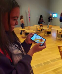 Student smiling at a photo on her phone of a scale model from "Sonya Clark: We Are Each Other", while standing in the same exhibition.