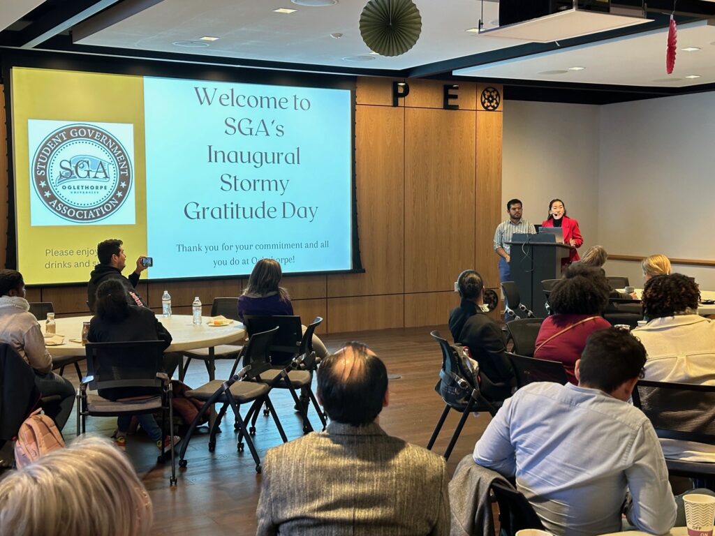 Ashleigh Ewald and David Martinez speaking at a podium to attendees with a powerpoint screen that reads "Welcome to SGA's Inaugural Stormy Gratitude Day. Thank you for your commitment and all you do at Oglethorpe!"
