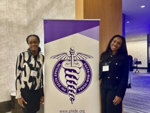 Camila Mondesir and Keturah Mba at the international convention in business casual wear standing next to a banner with the Phi Delta Epsilon logo.
